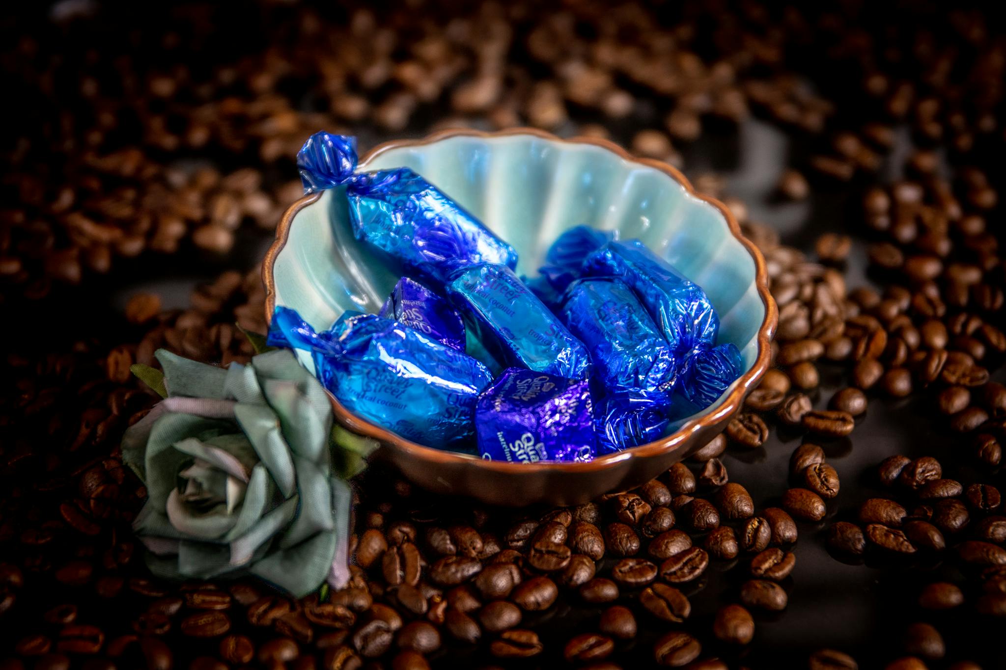 Close-up of blue wrapped chocolates in a ceramic bowl surrounded by coffee beans and a flower.