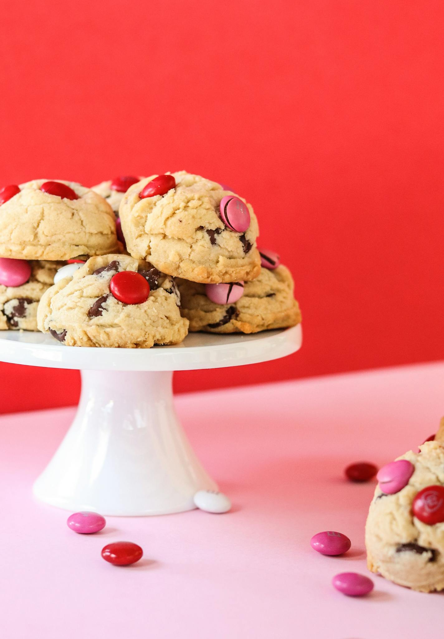 Stack of chocolate chip cookies with pink and red candy on a white cake stand. Vibrant and sweet.
